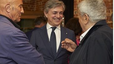 Three men converse at a social event; the man in the center wears a dark suit and tie, smiling as he talks with two others against a brick-walled background.