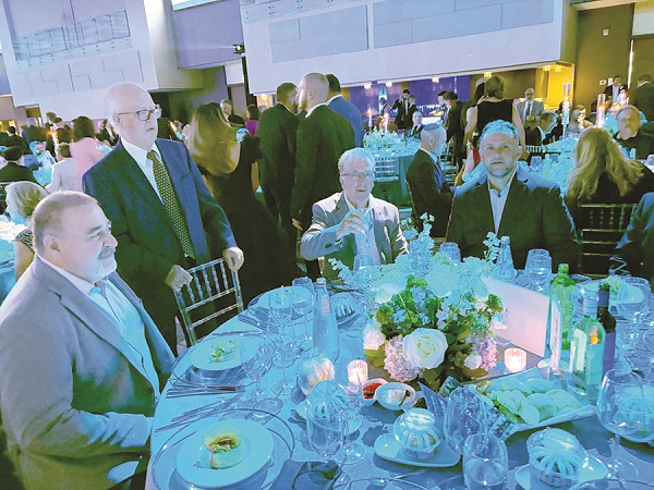 Formal dinner party with men in suits seated around a round table, floral centerpiece and candles on the table.