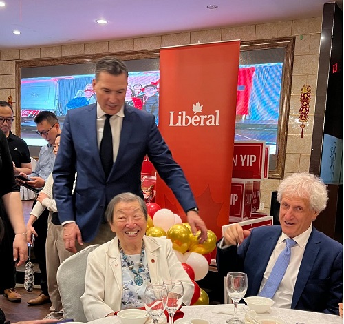 Smiling elderly woman in a white jacket sits at a banquet table during a Liberal party event, a man in a blue suit stands beside her with a red Liberal banner in the background.