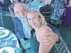 Couple seated at a banquet table, the man in a blue suit hugs the smiling woman in a black dress.