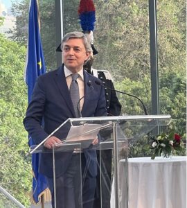 Man in a navy suit and tie speaks at a clear acrylic podium during a formal event, with microphones and a flag in the background.