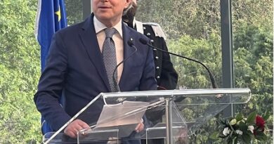 Man in a navy suit and tie speaks at a clear acrylic podium during a formal event, with microphones and a flag in the background.