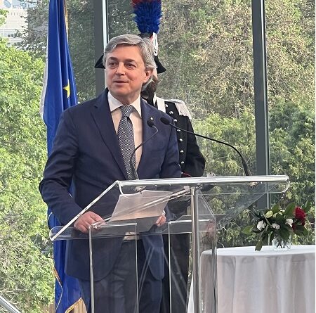 Man in a navy suit and tie speaks at a clear acrylic podium during a formal event, with microphones and a flag in the background.