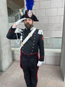 Man in ceremonial military dress uniform saluting, blue plume on hat, epaulettes, white gloves.