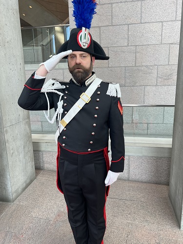 Man in ceremonial military dress uniform saluting, blue plume on hat, epaulettes, white gloves.