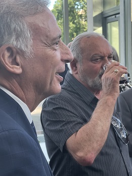 Profile of a man in a blue suit speaking, with another man in a striped shirt in the background smoking a cigarette outdoors.