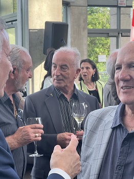 Group of older men in suits at an outdoor reception, holding wine glasses and chatting.