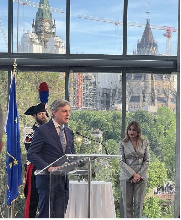 Man in a blue suit speaks at a podium while a woman in a gray suit stands nearby; EU flag and a city skyline are visible through large windows behind them.