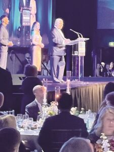 Man in a suit speaks at a clear lectern on stage during a formal event, with two people standing behind him on a lit backdrop.