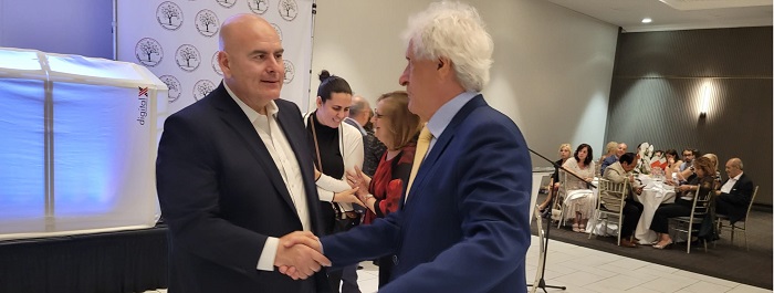 Two men in suits shake hands at a formal event, with guests seated at tables in the background.