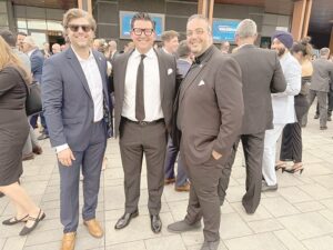 Three men in suits posing for a photo at a formal event, with a crowd behind them.