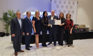 Group of seven professionals in formal attire posing as one person receives a certificate during a ceremony, backdrop with logos behind them.