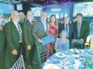 Group of eight adults in formal wear posing for a photo at a banquet; a seated elderly woman in a colorful dress sits at the front, with a Vaughan stage backdrop in the background.