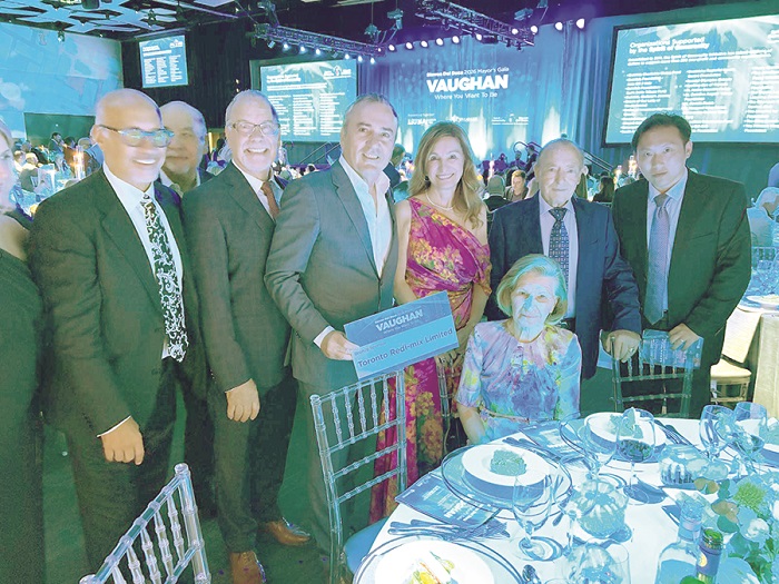 Group of eight adults in formal wear posing for a photo at a banquet; a seated elderly woman in a colorful dress sits at the front, with a Vaughan stage backdrop in the background.