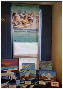 Colorful landscape paintings arranged on a table in a school hallway, with an Erasmus poster above.