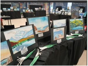 Rows of landscape paintings mounted on black display boards arranged on tables at an art exhibit in a gallery.