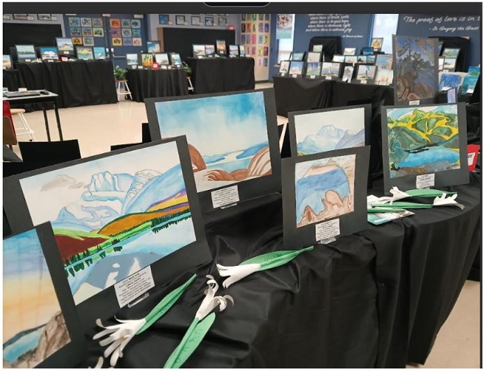 Rows of landscape paintings mounted on black display boards arranged on tables at an art exhibit in a gallery.