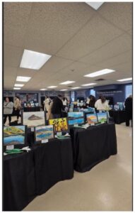Indoor art exhibit with several landscape paintings on black-cloth tables; attendees browse in background.