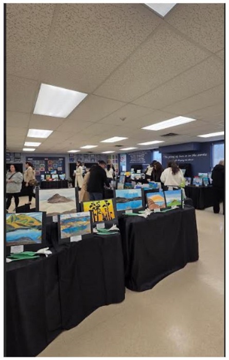 Indoor art exhibit with several landscape paintings on black-cloth tables; attendees browse in background.