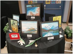 Student art exhibit: landscape paintings displayed on black mats around a round table; a Grade 7 sign sits beside white flower props.