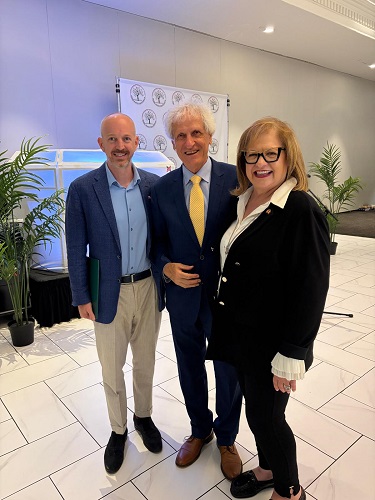 Three professionals pose together in a bright lobby; center man wears a blue suit and yellow tie, flanked by a bald man and a smiling woman.