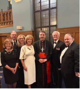 Group of seven adults in formal attire posing for a photo in a church hall; center figure is a clergyman in black robes with red trim.