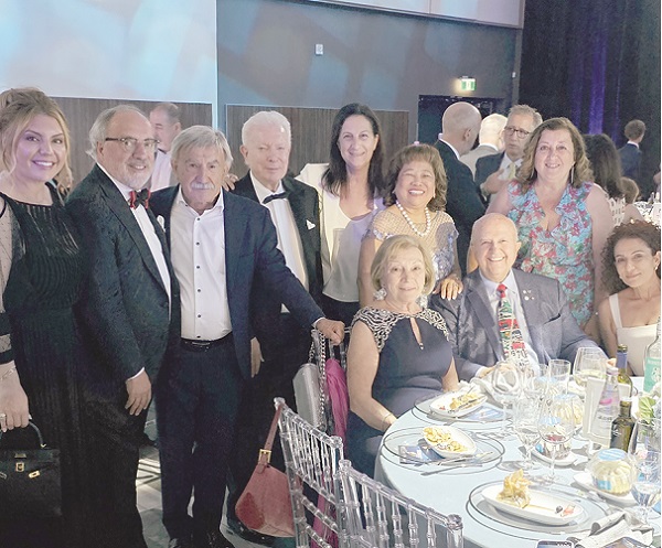 Group of formally dressed people posing for a photo at a round banquet table. They are at a formal event, with plates and glasses on the table and others standing in the background.