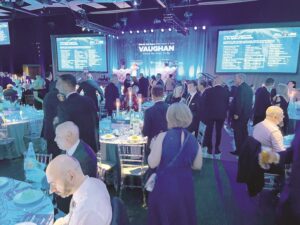 Formal gala or awards event in a large venue, round tables with teal tablecloths and candles, a blue-lit stage backdrop with screens.