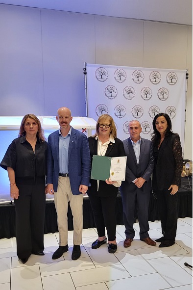 Five professionals standing in a row; the center woman holds a green binder and certificate, with a backdrop of circular logos behind them.