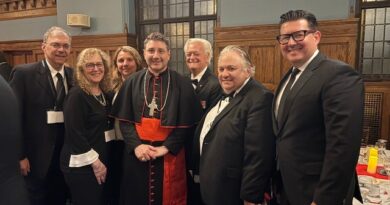 Eight people in formal attire posing for a group photo in a wood-paneled room; center figure is a clergyman in black and red robes with a cross, flanked by men in suits and bow ties.
