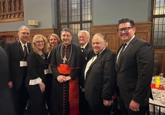 Eight people in formal attire posing for a group photo in a wood-paneled room; center figure is a clergyman in black and red robes with a cross, flanked by men in suits and bow ties.