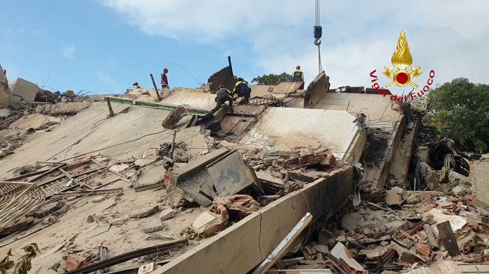 Firefighters conduct a rescue amid collapsed building debris on a rubble slope, with a crane overhead.