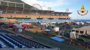 Stadium field filled with camping tents for emergency relief, ferris wheel in the background, blue arena seats nearby, and the Italian Fire Department emblem in the corner.