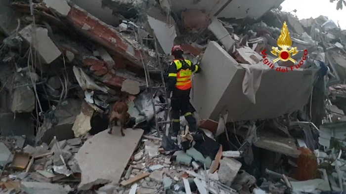 Firefighter with a helmet and high-visibility vest searching through rubble with a search dog; Vigili del Fuoco emblem visible