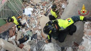 Italian Fire Department rescuers in bright yellow gear and helmets search through rubble and debris in a collapsed building.