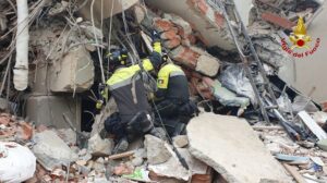 Firefighters in high-visibility gear perform a search-and-rescue amid collapsed building rubble, with bricks, concrete and tangled cables; Vigili del Fuoco emblem in the corner.