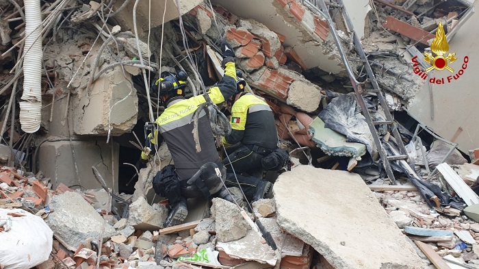 Firefighters in high-visibility gear perform a search-and-rescue amid collapsed building rubble, with bricks, concrete and tangled cables; Vigili del Fuoco emblem in the corner.