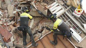 Two firefighters in yellow jackets search through a collapsed building amid rubble and debris under a partially standing facade.