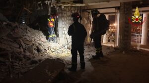 Firefighters in protective suits search through rubble inside a collapsed building at night with flashlights.