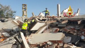 Firefighters in high-visibility jackets stand on and around a collapsed concrete building, inspecting rubble and securing a rope during a rescue operation.
