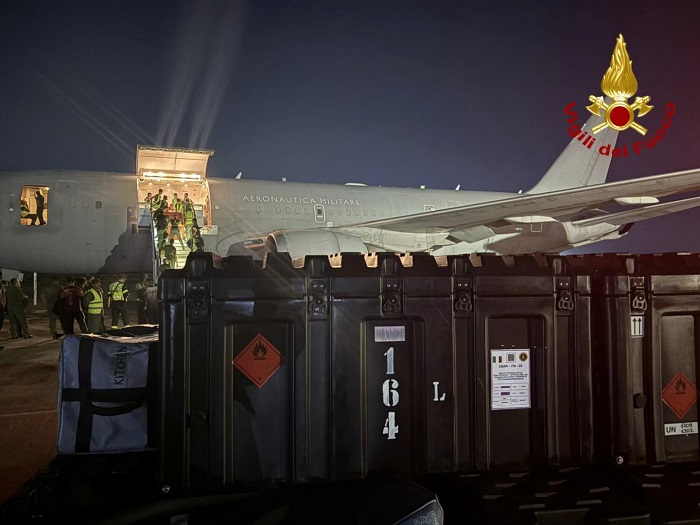 Night scene of a military cargo aircraft being unloaded at a tarmac, with large black crates and workers nearby; Italian fire service emblem visible in the corner.