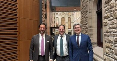 Three businessmen in suits pose together in a modern stone-walled hallway with a glass skylight above and wooden panels on the left.