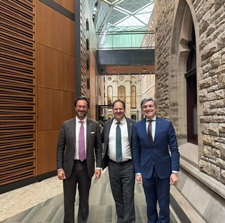 Three businessmen in suits pose together in a modern stone-walled hallway with a glass skylight above and wooden panels on the left.