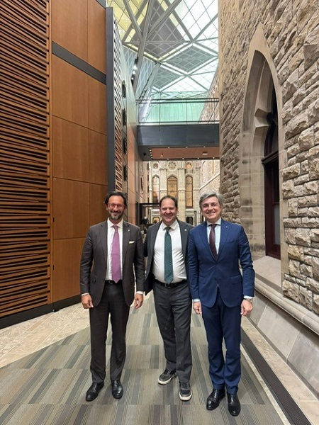 Three businessmen in suits pose together in a modern stone-walled hallway with a glass skylight above and wooden panels on the left.