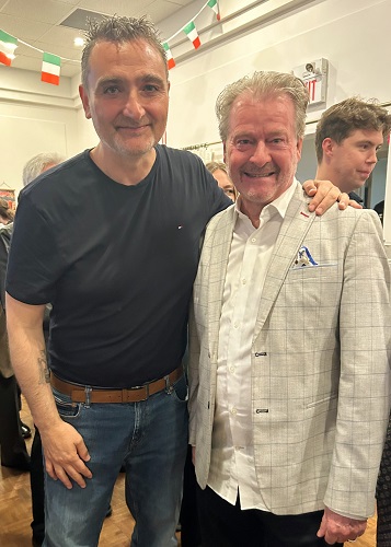 Two men posing for a photo at an indoor event, both smiling; one wears a black t-shirt and jeans, the other a light gray check blazer with a white shirt.