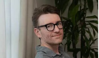 Man with short brown hair and black glasses sits indoors, wearing a gray polo and smiling slightly.