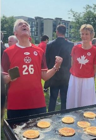 Man in red jersey #26 gestures triumphantly beside a pancake griddle as pancakes cook, with a woman in a red maple-leaf top looking on.