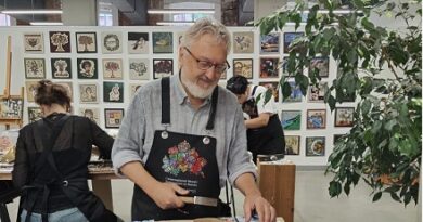 Smiling man with gray beard wearing a dark apron works on a ceramic piece at a pottery studio, with colorful mosaics and tools on the table nearby.