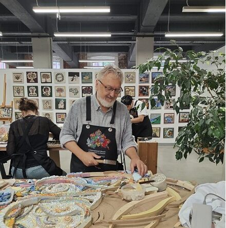 Smiling man with gray beard wearing a dark apron works on a ceramic piece at a pottery studio, with colorful mosaics and tools on the table nearby.