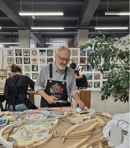 Smiling man with gray beard wearing a dark apron works on a ceramic piece at a pottery studio, with colorful mosaics and tools on the table nearby.